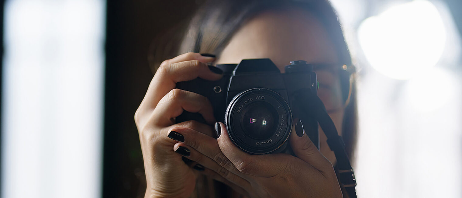 Fotografin im Fokus: Kamera vor dem Gesicht im natürlichen Licht Frau mit dunklem Nagellack fotografiert mit Spiegelreflexkamera im Gegenlicht