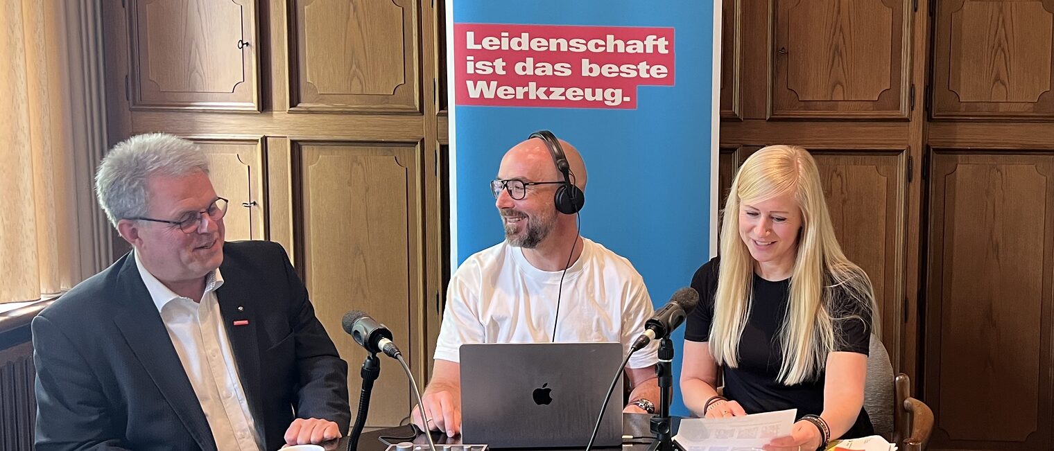Drei Personen bei einer Podcast-Aufzeichnung am Tisch mit Mikrofonen, Laptop Roll-Up im Hintergrund.