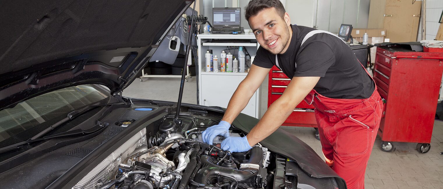Junger Kraftfahrzeugmechatroniker in roter Arbeitshose lächelt beim Arbeiten am geöffneten Motorraum eines Autos