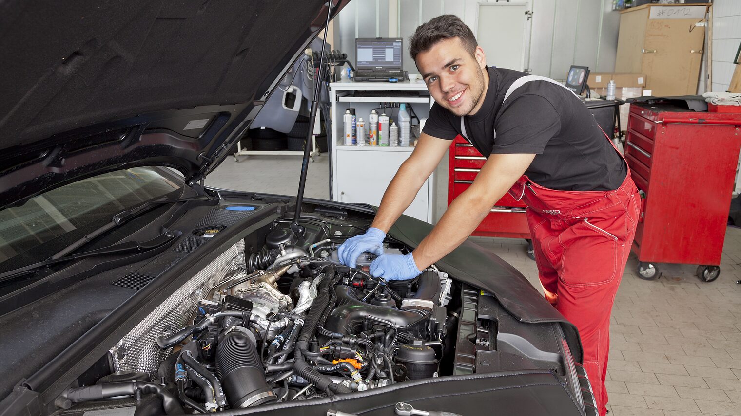 Junger Kraftfahrzeugmechatroniker in roter Arbeitshose lächelt beim Arbeiten am geöffneten Motorraum eines Autos